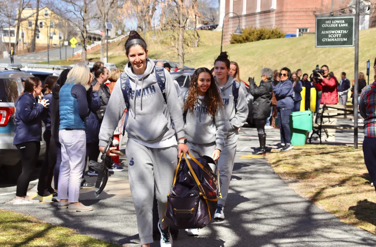 Basketball players, carrying luggage and smiling, walk along the path toward the bus