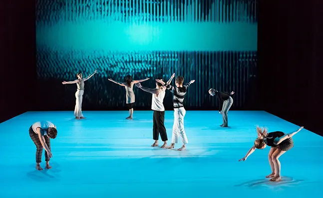 Seven dancers on stage against a bright blue floor and backdrop
