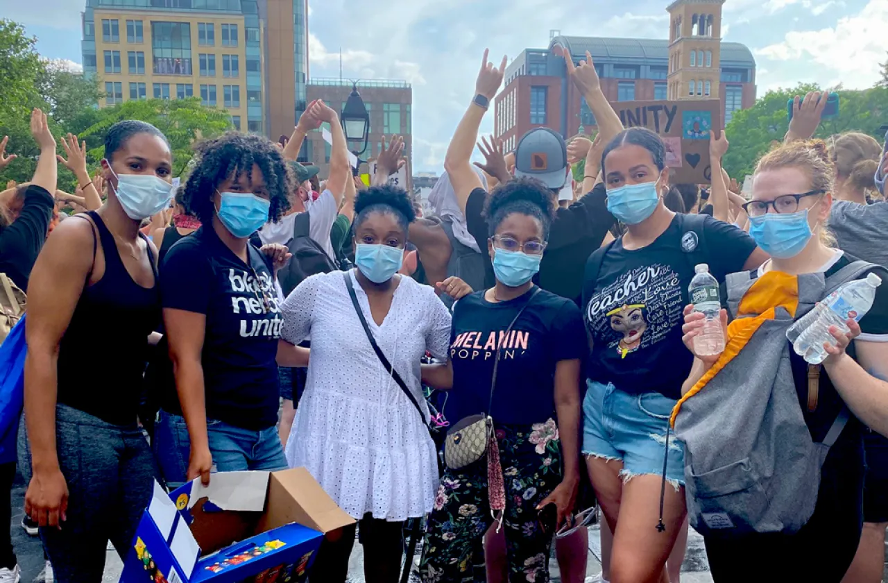 Six friends—five black and one white— in masks standing next to each other in front of a citscape. T-shirts include "Black Nerds Unite" and "Melanin Poppin"