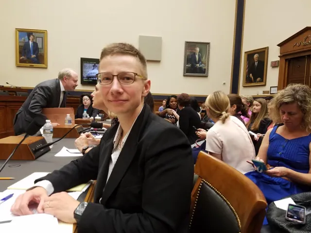 Aleks Kajstura in a suit with short hair at a desk in what looks like a government hall with others in the background