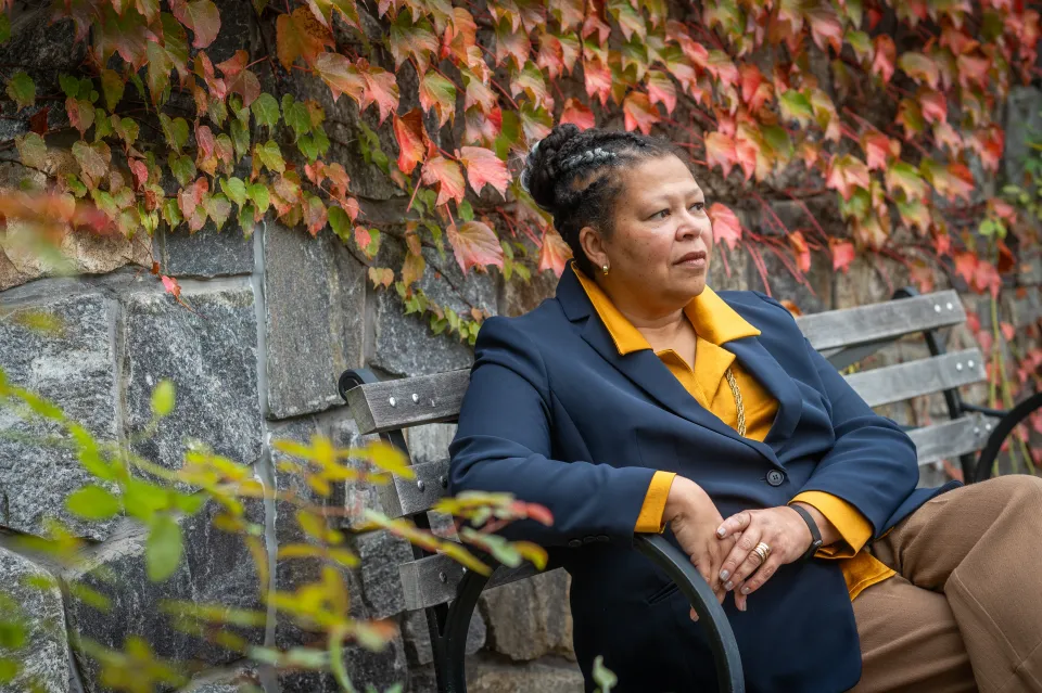 Photo of Sarah Willie-LeBreton sitting on a bench and looking into the distance, with fall foilia