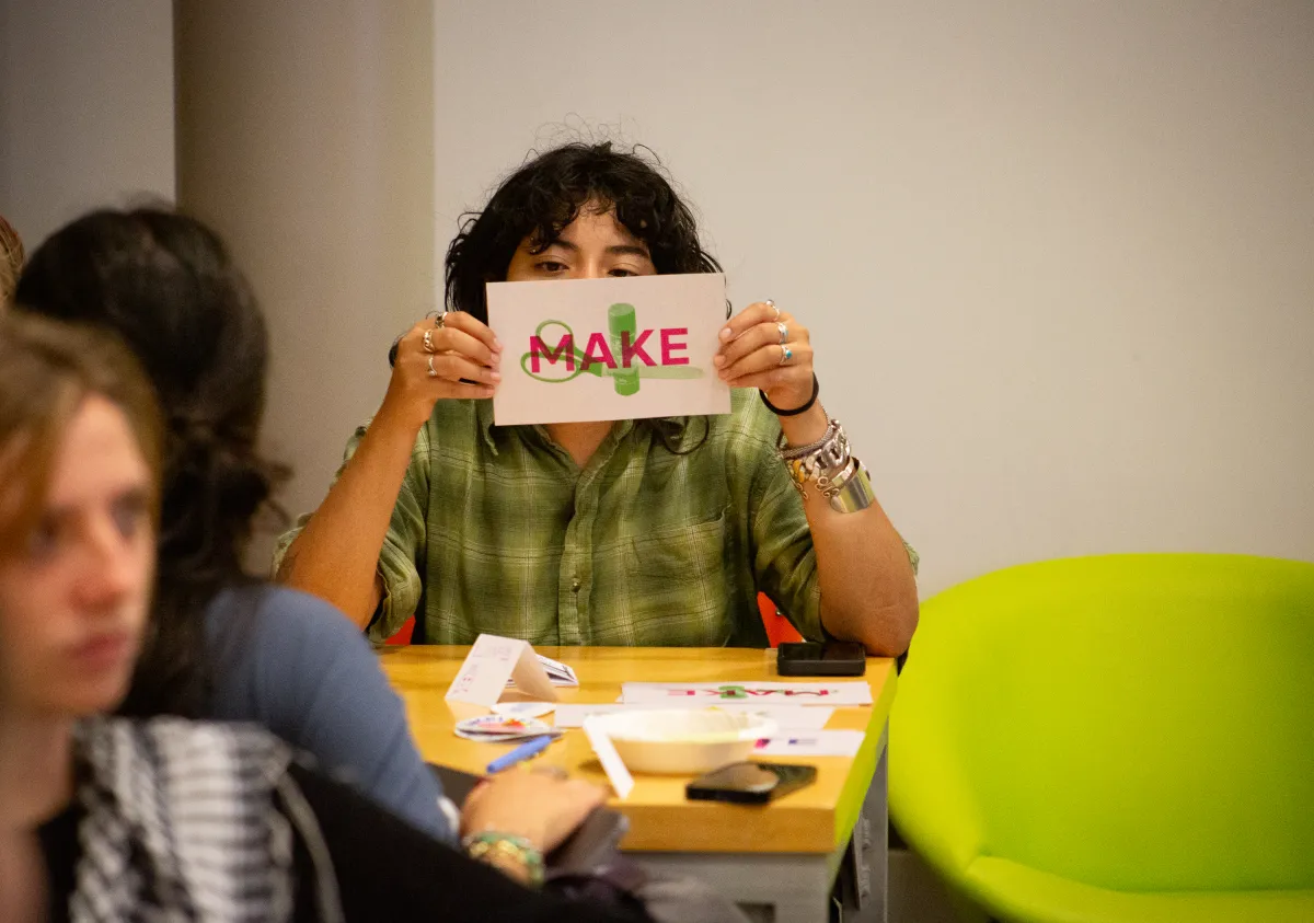 A student holds a paper with the words Make in front of their face while looking at the camera at ZineFest 2024