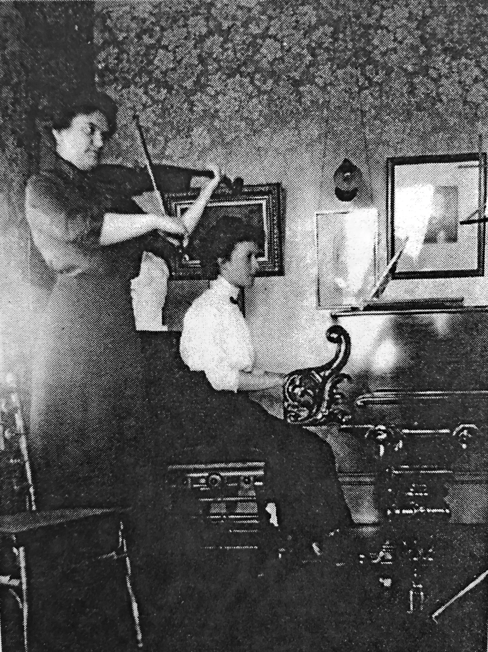 Antique photo of two women playing music — one standing playing the violin and the other sitting playing the piano.