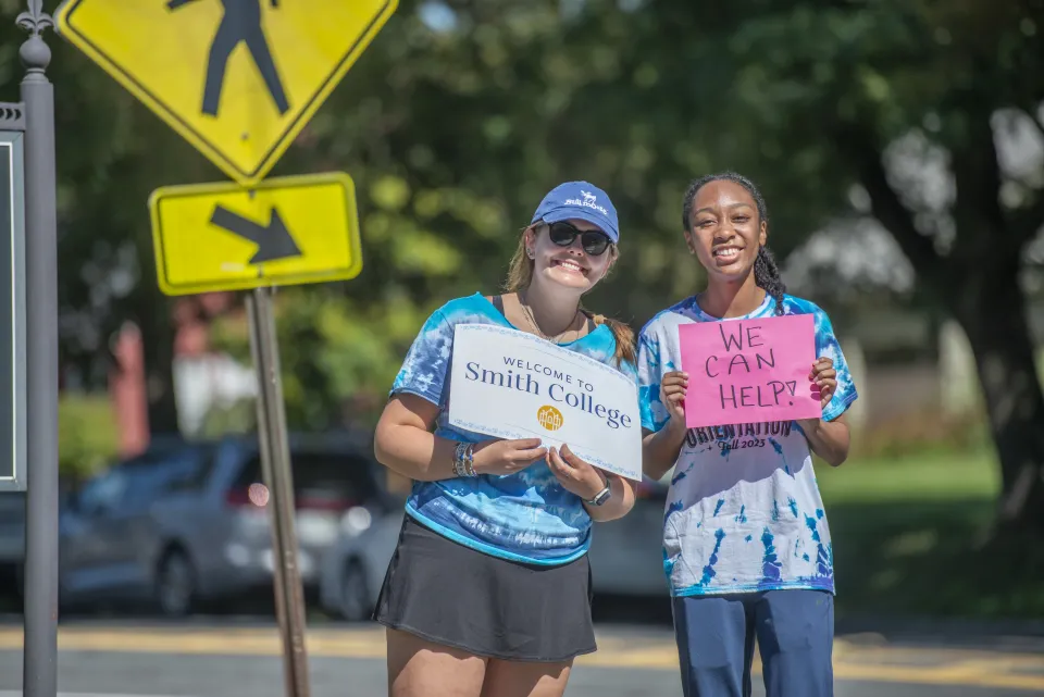 Two students holding signs that say "Welcome to Smith College" and "We can help!" on move-in day