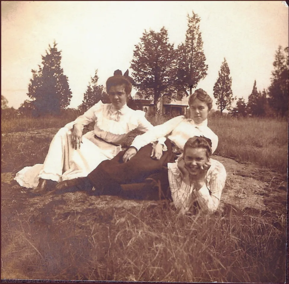 An antique photo of three women reclining and posing in an open field.