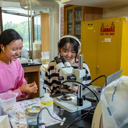 Two students, one in pink and one in stripes with headphones and earbuds, work at microscopes at Professor Marney Pratt's lab in Sabin-Reed